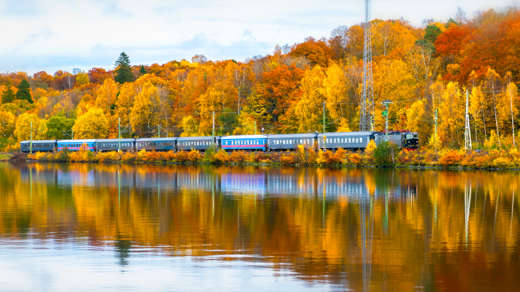 Ett blått tåg rullar längs sjökant med höstfärgad färgglad skog.