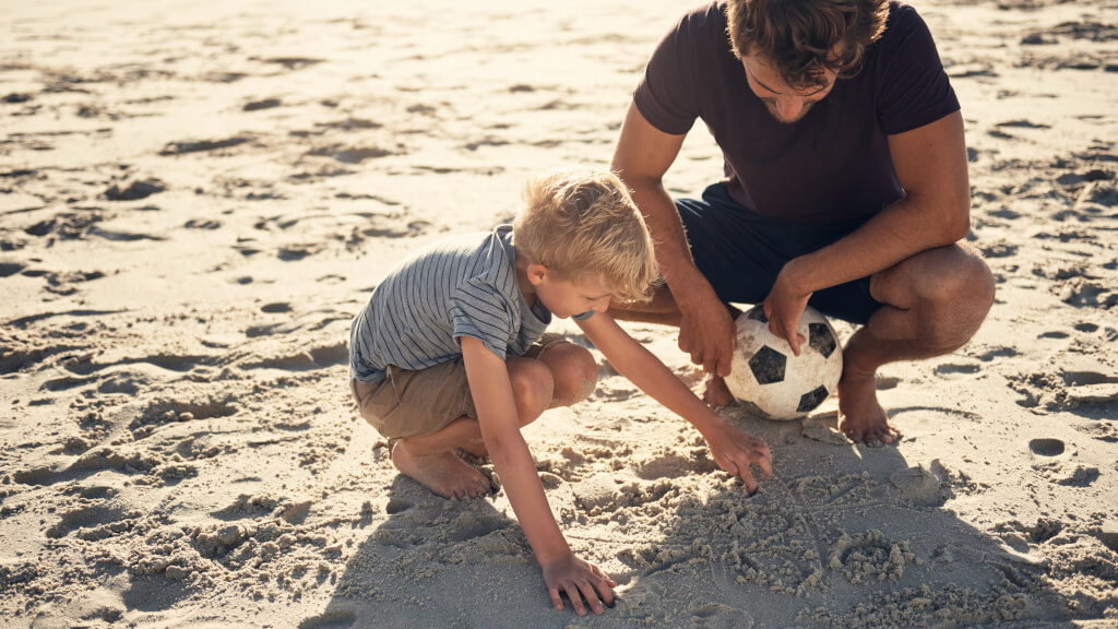 Pappa med fotboll och pojke sitter på sandstrand och ritar i sanden tillsammans.