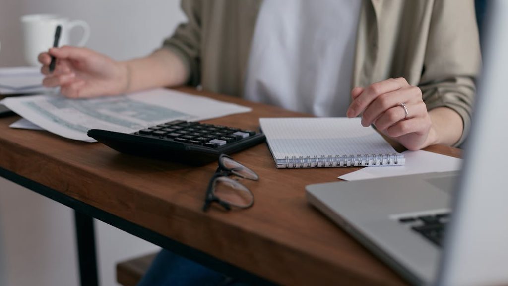 A woman manages finances at home, using a laptop and calculator on a wooden desk.